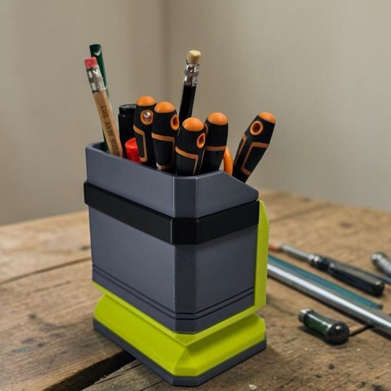 May include: A grey and neon green desk tidy holding pencils and screwdrivers. The organiser has a rectangular shape with a black band around the middle. The base is neon green, and the top section is grey.