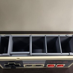 May include: A gray and cream-colored organizer with multiple compartments, resembling a classic video game controller. The design includes a directional pad, buttons, and the text "SELECT START" and "CHARLIE". The compartments are divided by gray dividers.