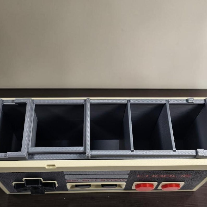 May include: A gray and cream-colored organizer with multiple compartments, resembling a classic video game controller. The design includes a directional pad, buttons, and the text "SELECT START" and "CHARLIE". The compartments are divided by gray dividers.