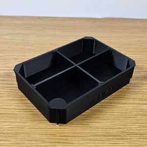 Puede incluir: Un organizador rectangular negro con cuatro compartimentos. El organizador est&aacute; hecho de un material texturizado y tiene una esquina redondeada. El texto "CUBE-ID" est&aacute; impreso en el lateral.