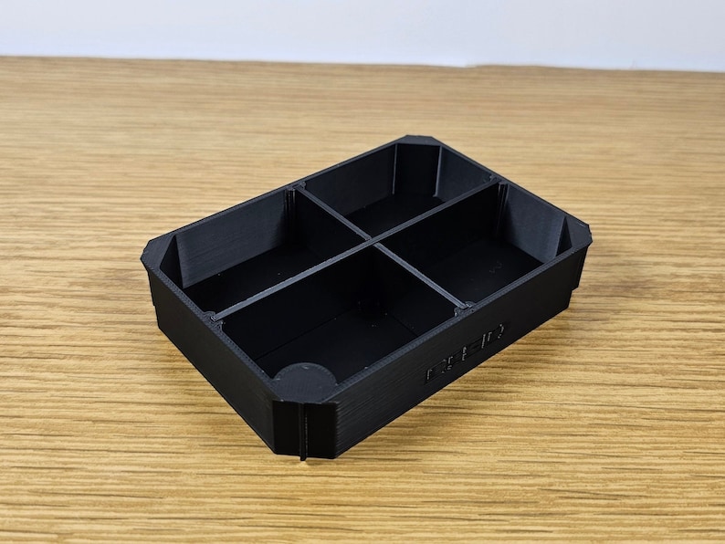 Puede incluir: Un organizador rectangular negro con cuatro compartimentos. El organizador est&aacute; hecho de un material texturizado y tiene una esquina redondeada. El texto "CUBE-ID" est&aacute; impreso en el lateral.
