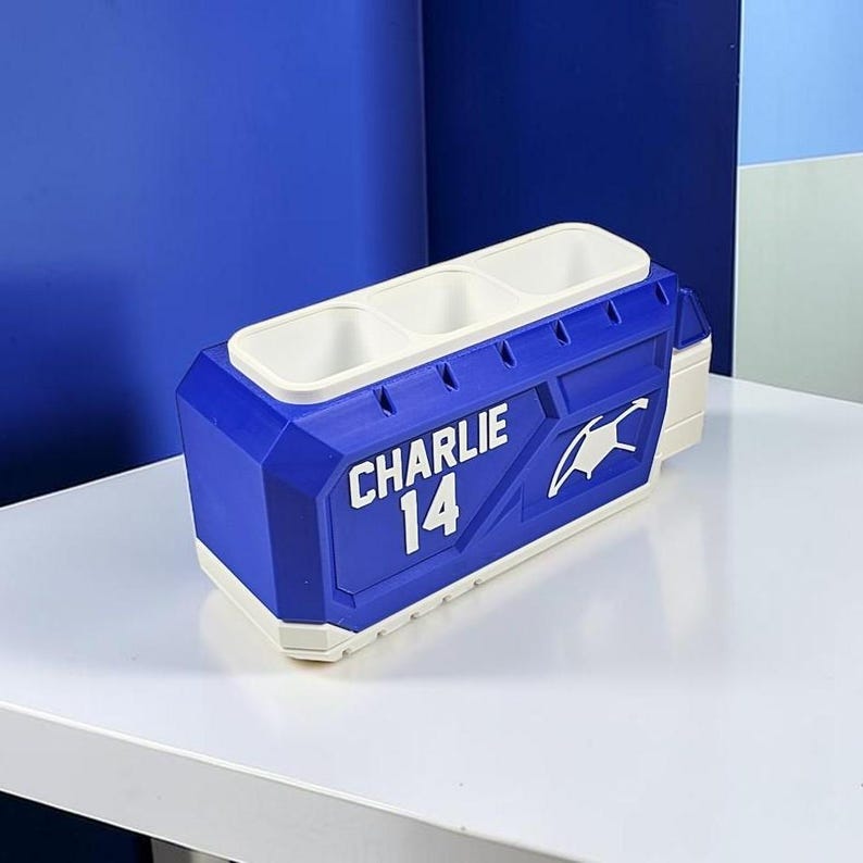 May include: A blue and white rectangular desk organizer with three top compartments. The side features the white text CHARLIE and the number 14, along with a white graphic icon of a sports jersey. The base and top rim are white.