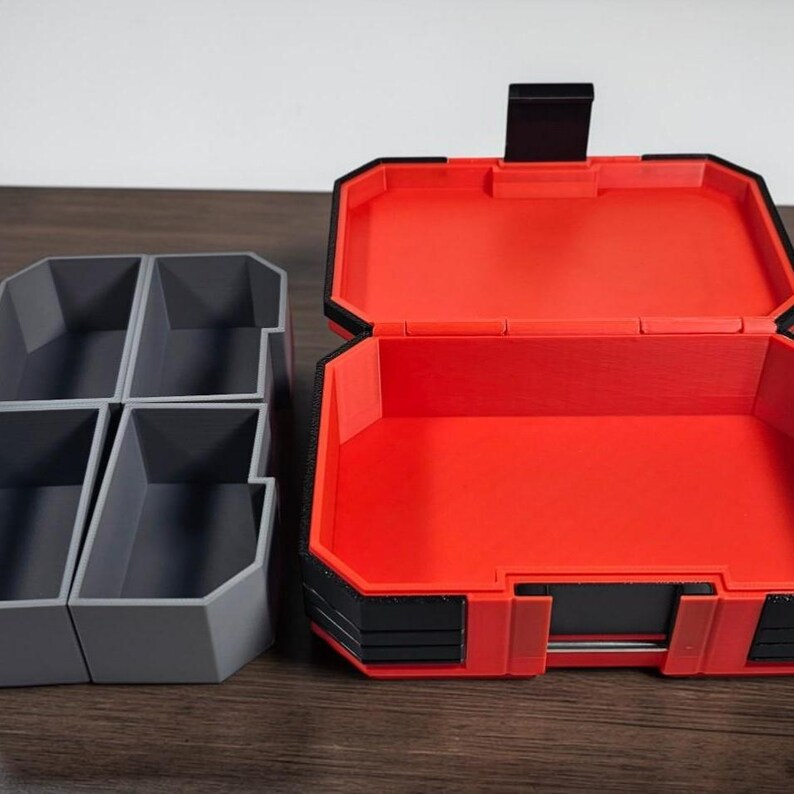 May include: A red and black toolbox with an open lid, revealing an empty interior. To the left, there are four grey storage containers with individual compartments. The items are on a wooden surface.