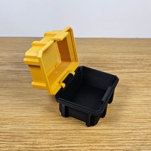 May include: A small yellow plastic storage box with a black lid. The lid is open and the box is empty.