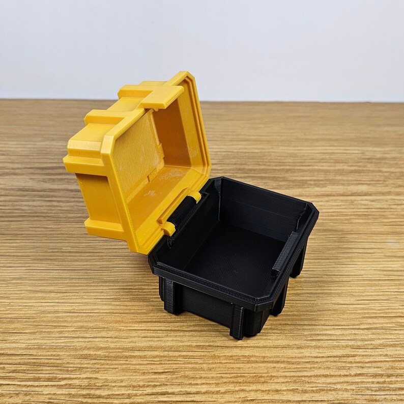 May include: A small yellow plastic storage box with a black lid. The lid is open and the box is empty.
