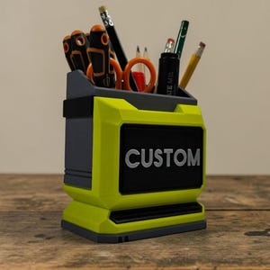 May include: A two-tone desk organiser in grey and neon yellow, with the word "CUSTOM" in white lettering on a black panel. It contains office supplies such as pencils, scissors, and screwdrivers. The organiser sits on a wooden surface.