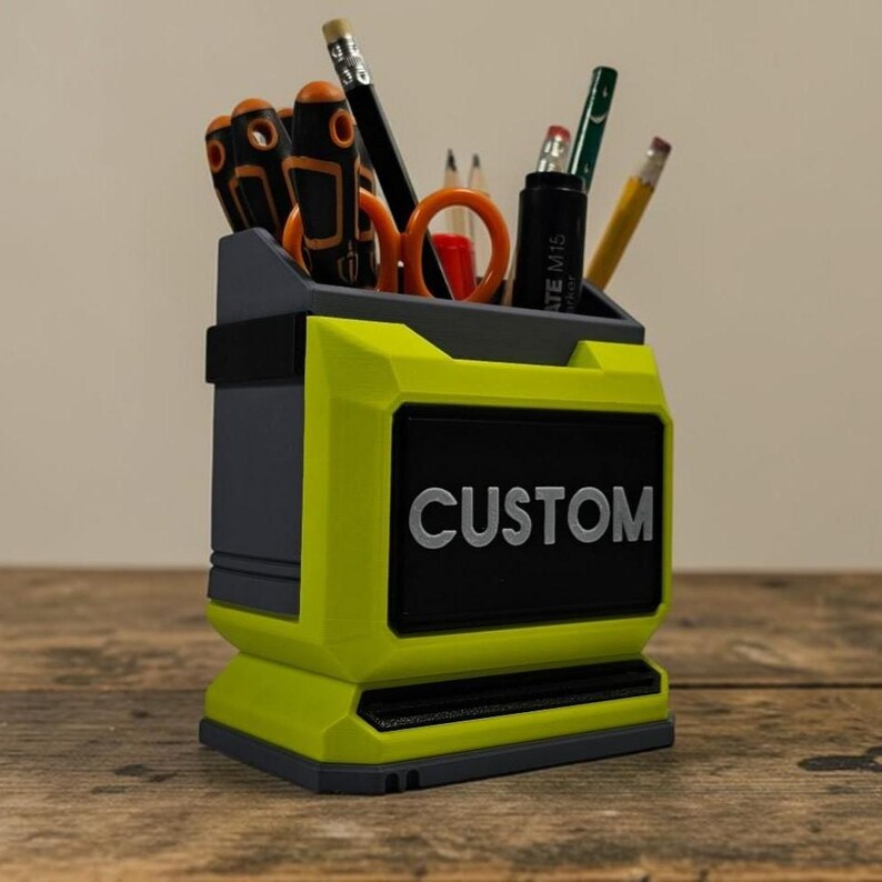May include: A two-tone desk organiser in grey and neon yellow, with the word "CUSTOM" in white lettering on a black panel. It contains office supplies such as pencils, scissors, and screwdrivers. The organiser sits on a wooden surface.
