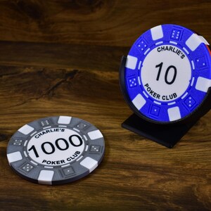 Może przedstawiać: Dwa żetony do pokera z napisem "Charlie's Poker Club" i numerami "10" i "1000". Jeden żeton jest niebiesko-biały, drugi szaro-biały. Niebieski żeton jest w czarnym stojaku. Żetony leżą na drewnianej powierzchni.