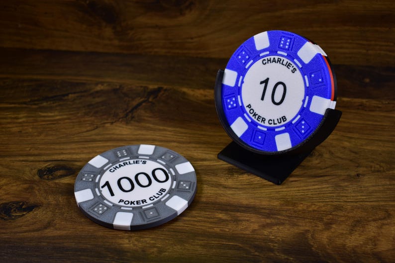 Może przedstawiać: Dwa żetony do pokera z napisem "Charlie's Poker Club" i numerami "10" i "1000". Jeden żeton jest niebiesko-biały, drugi szaro-biały. Niebieski żeton jest w czarnym stojaku. Żetony leżą na drewnianej powierzchni.