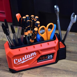 May include: A custom-designed orange and black tool organizer with the word "Custom" and the number "1S425" printed on the front. The organizer holds pens, screwdrivers, scissors, and wrenches. The tools are neatly arranged inside the organizer.