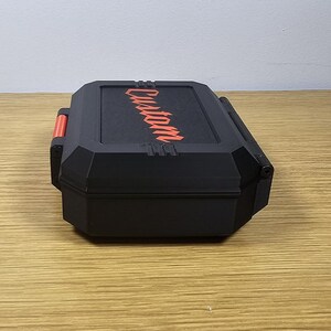 May include: Black rectangular case with the word "Custom" in red diagonally across the top. The case has a red accent on the side and is sitting on a wooden surface. The case appears to be a storage container.