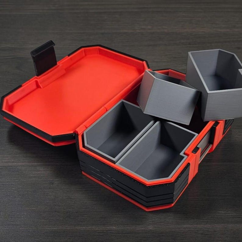 May include: Open, red and black storage box with grey interior compartments. The box has a hinged lid and several removable grey containers. The box is designed with a geometric, angular shape.