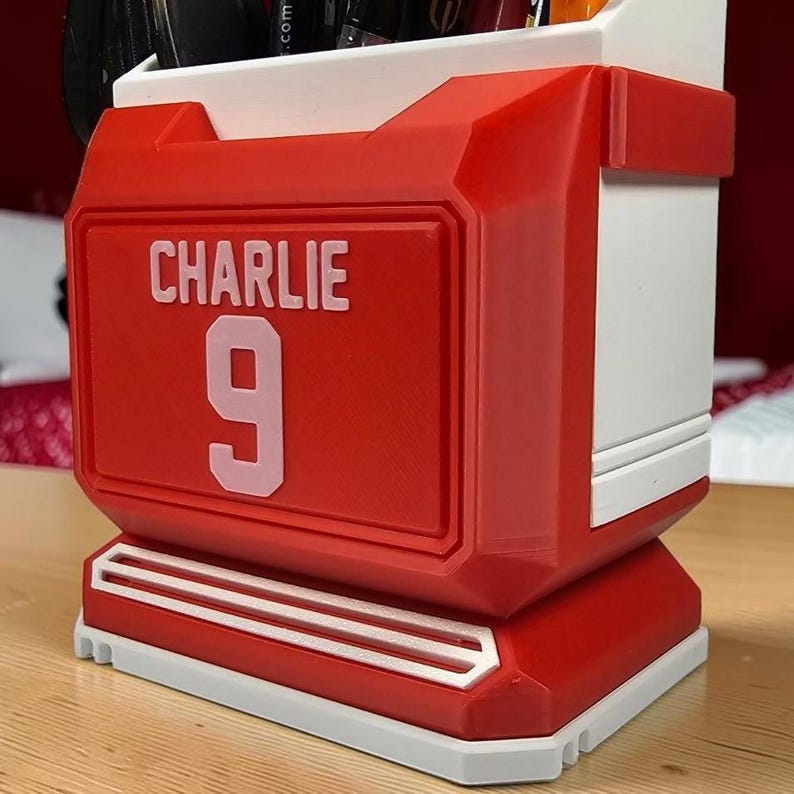 May include: A red and white desk organizer shaped like a sports jersey, featuring the name CHARLIE and the number 9 in white lettering on the front. The container has a rectangular structure with a flat base and an open top for holding pens.