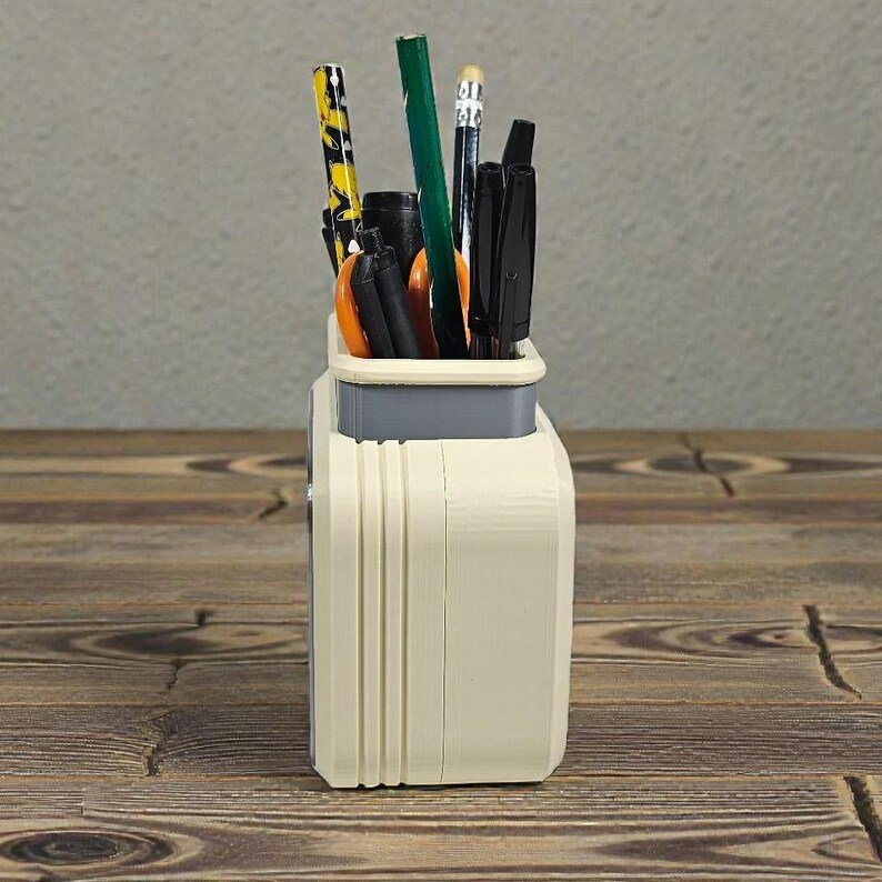 May include: A cream-colored desk organizer with a gray top section, holding various pens, pencils, and scissors. The organizer has vertical ridged detailing on the front and sits on a wooden surface.
