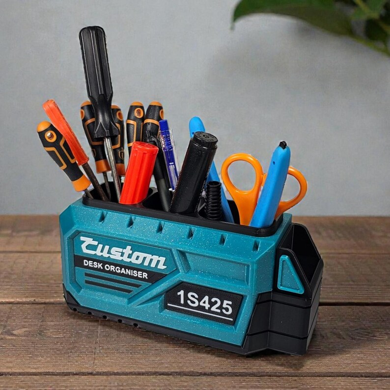 May include: A teal and black desk organizer with the word "Custom" and "DESK ORGANISER" printed on the front. The organizer holds various tools, including screwdrivers, pens, and scissors. The model number "1S425" is also printed on the front.