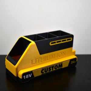 Puede incluir: Un organizador de escritorio amarillo y negro con forma de bater&iacute;a, con el texto "LITHIUM ION CUSTOM" y "18V". El organizador tiene cuatro compartimentos y un frente inclinado.
