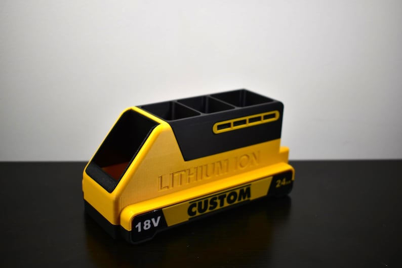 Puede incluir: Un organizador de escritorio amarillo y negro con forma de bater&iacute;a, con el texto "LITHIUM ION CUSTOM" y "18V". El organizador tiene cuatro compartimentos y un frente inclinado.