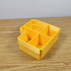 May include: A bright yellow, multi-compartment organizer. The rectangular container has a stepped design with five distinct sections. The organizer is made of a solid, smooth material and is designed for storage and organization.