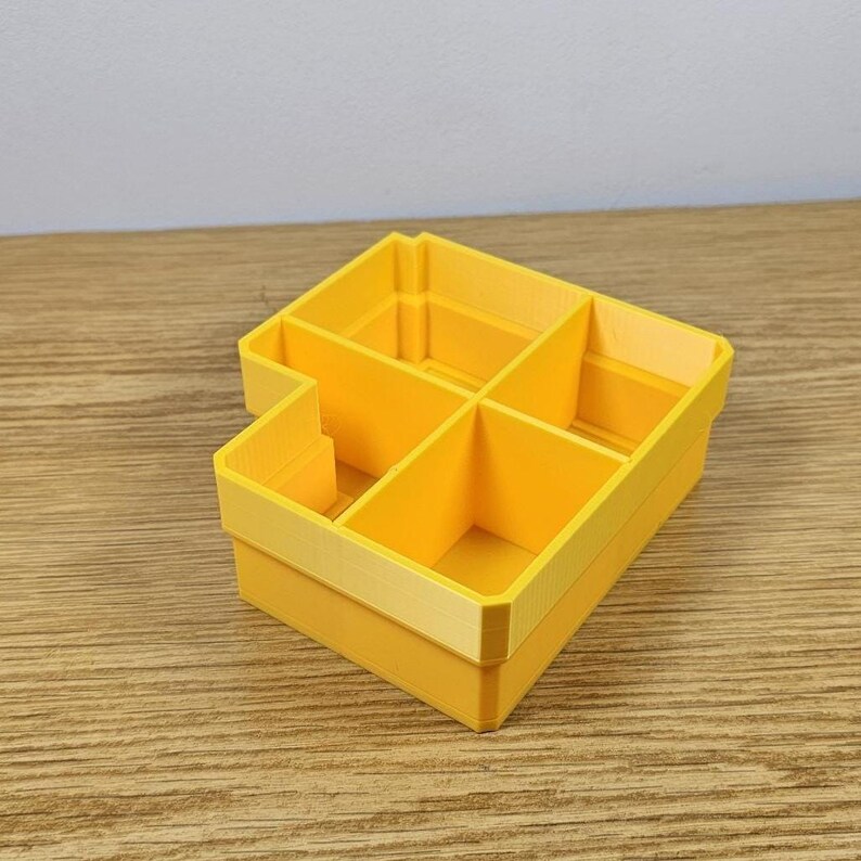 May include: A bright yellow, multi-compartment organizer. The rectangular container has a stepped design with five distinct sections. The organizer is made of a solid, smooth material and is designed for storage and organization.