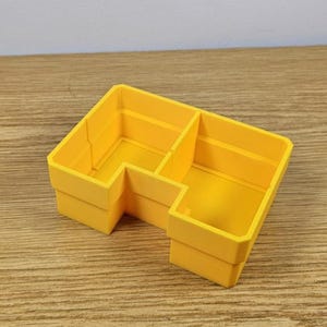 May include: A bright yellow, L-shaped organizer with three compartments. The organizer is made of a solid material and sits on a wooden surface. The compartments are divided by vertical walls, creating separate storage spaces.