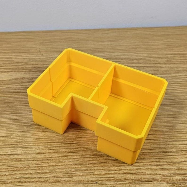 May include: A bright yellow, L-shaped organizer with three compartments. The organizer is made of a solid material and sits on a wooden surface. The compartments are divided by vertical walls, creating separate storage spaces.