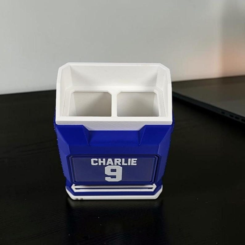 May include: A blue and white personalized pencil holder with the name "CHARLIE" and the number "9" on the front. The holder has two compartments and is made of plastic.