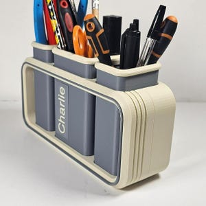 May include: A three-compartment desk organizer in a cream and gray color scheme. The organizer holds various pens, pencils, and scissors. The name "Charlie" is printed on the front. The design features rounded edges and a modern aesthetic.