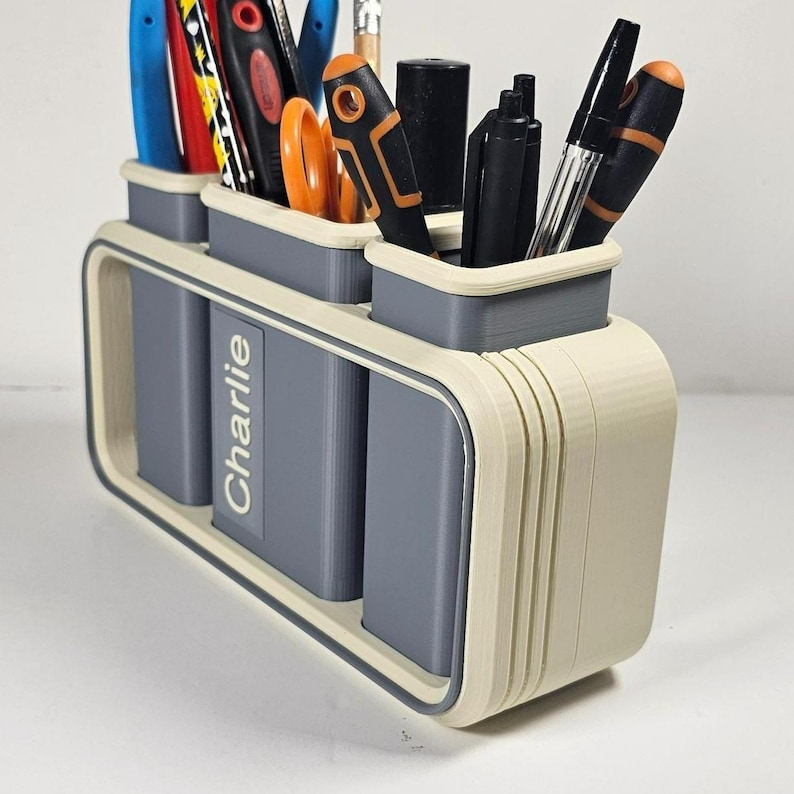 May include: A three-compartment desk organizer in a cream and gray color scheme. The organizer holds various pens, pencils, and scissors. The name "Charlie" is printed on the front. The design features rounded edges and a modern aesthetic.