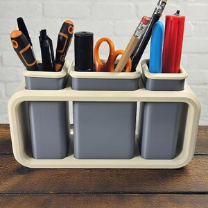 May include: A desk organizer with a cream-colored frame and gray containers. The organizer holds various office supplies, including pens, pencils, scissors, and markers. The desk organizer is sitting on a wooden surface.
