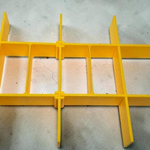 May include: Bright yellow plastic grid structure with intersecting horizontal and vertical bars, creating rectangular compartments. The grid is designed for organization and storage, with a simple, functional aesthetic.