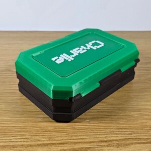 May include: A small, green and black box with the word "ALIEN" printed on the top. The box is made of plastic and has a hinged lid.