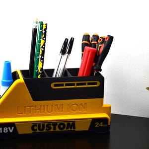 Puede incluir: Un organizador de escritorio amarillo y negro con forma de bater&iacute;a, que contiene bol&iacute;grafos, l&aacute;pices y destornilladores. El organizador tiene las palabras "LITHIUM ION" y "CUSTOM" impresas. Tambi&eacute;n se ve un bote de pegamento.