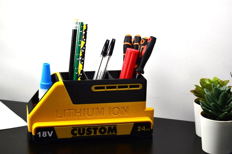 Puede incluir: Un organizador de escritorio amarillo y negro con forma de bater&iacute;a, que contiene bol&iacute;grafos, l&aacute;pices y destornilladores. El organizador tiene las palabras "LITHIUM ION" y "CUSTOM" impresas. Tambi&eacute;n se ve un bote de pegamento.