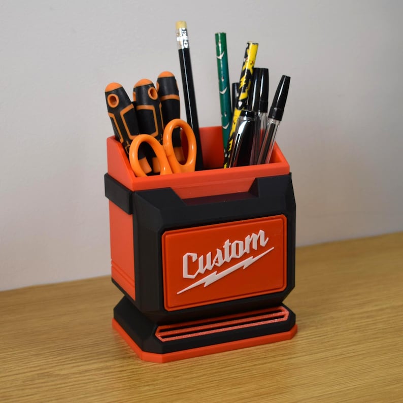 May include: A custom-designed desk organizer in black and orange, holding various writing utensils and small tools. The organizer features the word "Custom" in white lettering on the front. The design is reminiscent of a toolbox.
