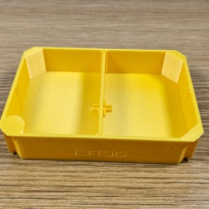 May include: A yellow plastic tray with two compartments. The tray has a raised edge around the perimeter and a small divider in the middle.