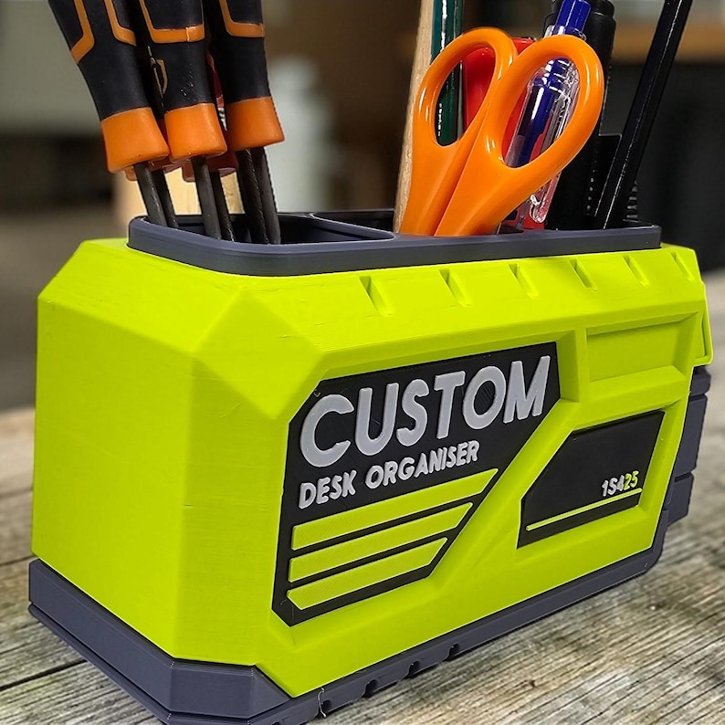 May include: A lime green desk organizer with a gray trim, holding tools and office supplies. The organizer has the word "CUSTOM DESK ORGANISER" printed on the front. The organizer contains orange scissors, pens, and screwdrivers.