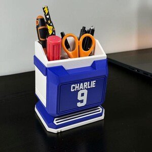 May include: A blue and white desk organizer with the name "CHARLIE" and the number "9" on the front. The organizer holds pens, scissors, and other office supplies. The design is modern and geometric.