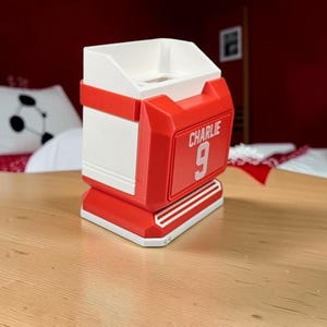 May include: A white and red plastic desk organizer shaped like a sports jersey display. The front features a red rectangular panel with the white text CHARLIE and the number 9 printed on it. The structure has a wide base and an open top compartment.