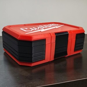 May include: A red and black tool box with the word "Custom" in white lettering and a white lightning bolt design on the lid. The box has a rectangular shape with a hinged lid and a series of black panels on the sides.