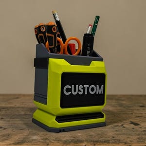 May include: A custom desk tidy in yellow and grey, with a black panel displaying "CUSTOM" in white. It contains tools such as screwdrivers, scissors, and pencils. This organiser is suitable for a desk or workshop, and is approximately 15 cm in height.