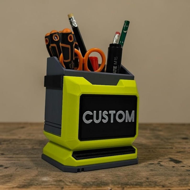 May include: A custom desk tidy in yellow and grey, with a black panel displaying "CUSTOM" in white. It contains tools such as screwdrivers, scissors, and pencils. This organiser is suitable for a desk or workshop, and is approximately 15 cm in height.