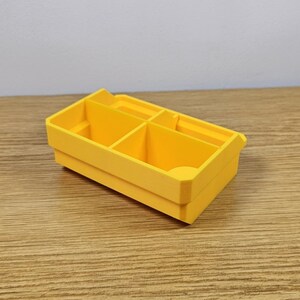 May include: A bright yellow, rectangular organizer with multiple compartments. The organizer is made of a solid material and sits on a wooden surface. The compartments are divided by vertical dividers, creating separate storage spaces.