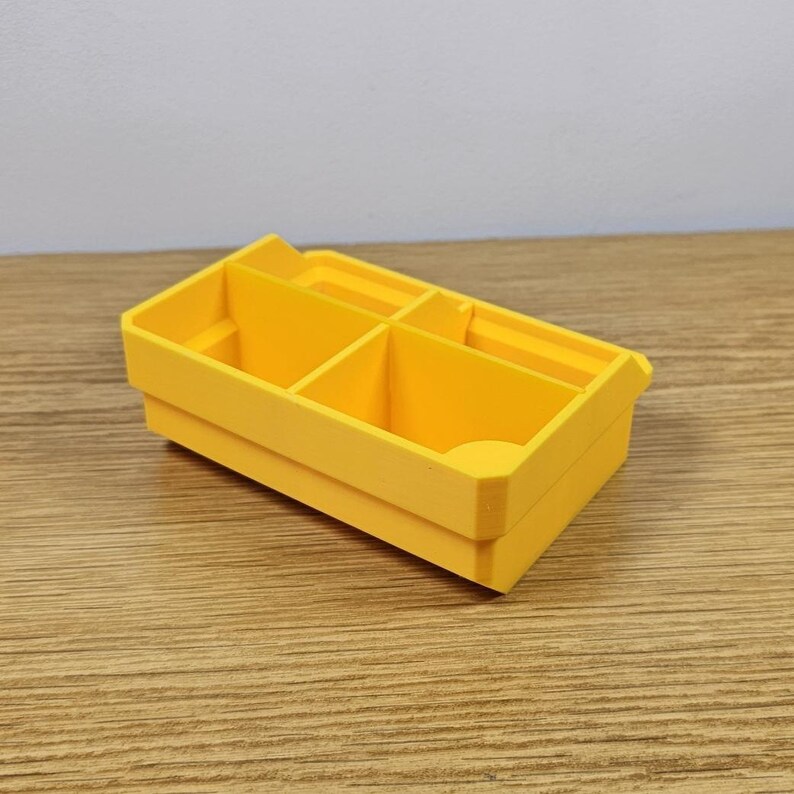 May include: A bright yellow, rectangular organizer with multiple compartments. The organizer is made of a solid material and sits on a wooden surface. The compartments are divided by vertical dividers, creating separate storage spaces.
