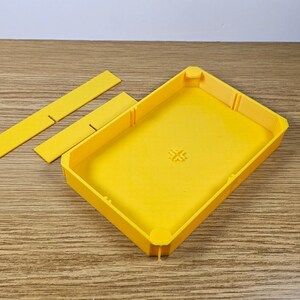 May include: A yellow plastic box with a rectangular shape and a small cross design on the top. The box is open and has three separate pieces that fit inside.