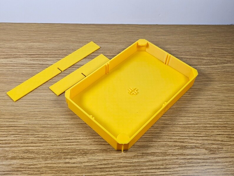 May include: A yellow plastic box with a rectangular shape and a small cross design on the top. The box is open and has three separate pieces that fit inside.