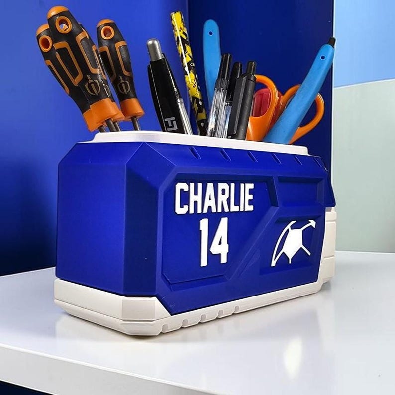 May include: A blue and white rectangular desk organizer featuring the name CHARLIE and the number 14 printed in white on the front. The container is filled with various office supplies including orange and black screwdrivers, pens, and scissors.
