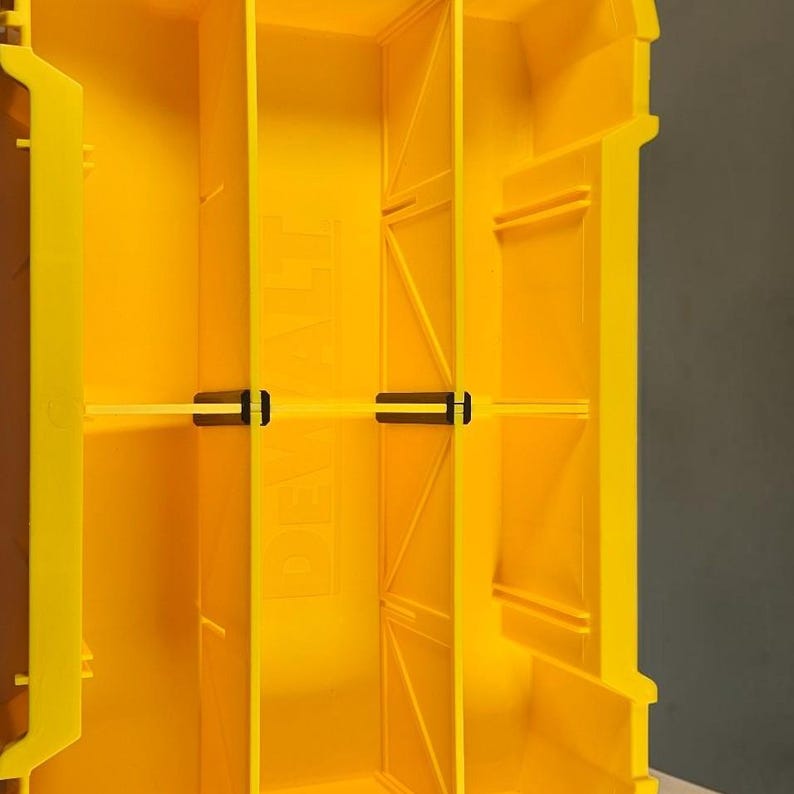 May include: A bright yellow plastic storage organizer featuring vertical dividers and a horizontal crossbar. The interior walls display the embossed text DEWALT. The structure includes molded support ribs and small black plastic clips.