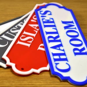 Puede incluir: Varios letreros decorativos en diferentes colores, incluyendo negro, rojo y blanco. Un letrero dice "CHARLIE'S ROOM" en letras azules. Los letreros tienen un borde decorativo y est&aacute;n dispuestos sobre una superficie de madera.