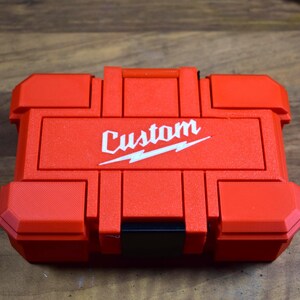 May include: A red, rectangular tool case with the word "Custom" in white script and a lightning bolt design. The case has a handle and multiple compartments, and is made of a durable plastic material.