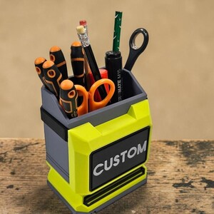 May include: A custom desk tidy in grey and neon yellow, containing tools and stationery. The organiser has the word "CUSTOM" in white on a black label. It holds screwdrivers, scissors, pencils, and a pen. The design resembles a toolbox.